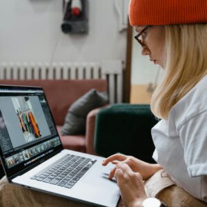 Young woman in orange knit hat browsing online on laptop indoors, showcasing fashion and technology.