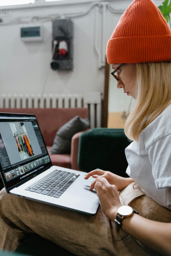 Young woman in orange knit hat browsing online on laptop indoors, showcasing fashion and technology.